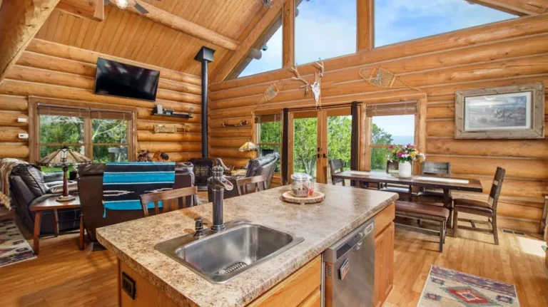 Cozy log cabin interior with an open-plan kitchen and living area featuring wooden furnishings and large windows.